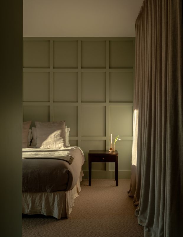 A softly lit, intimate bedroom with a partially visible upholstered bed dressed in layered neutral bedding, a dark wood bedside table, and a small vase. The room features a full-height grid-style wall panel treatment in a muted sage-grey tone, plush carpet, and tall, heavy pleated curtains filtering warm sunlight. The overall atmosphere is refined, calm, and cosy.