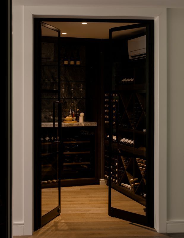 A dark, refined wine storage room viewed through open glass-and-black-framed double doors. The compact space contains extensive built-in wine racks and bottle storage, a stone-topped counter with a small sink or preparation area, dark feature walls, warm timber flooring, recessed ceiling lighting, and a wall-mounted cooling unit. White surrounding wall mouldings frame the entrance.