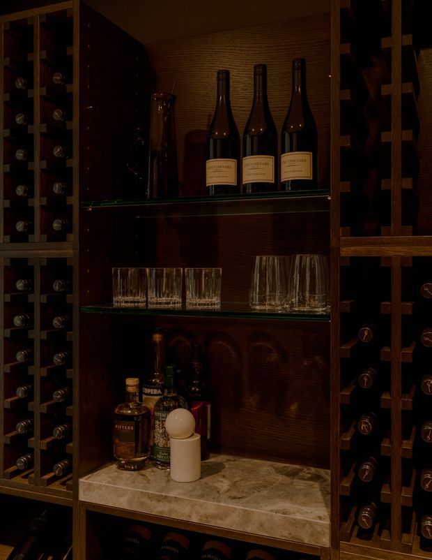 Dark, richly finished built-in wine storage with vertical bottle racks on both sides, central glass shelves displaying wine bottles and tumblers, and a marble-look counter below with spirits and a small lamp. The space has a moody, intimate cellar or bar atmosphere.