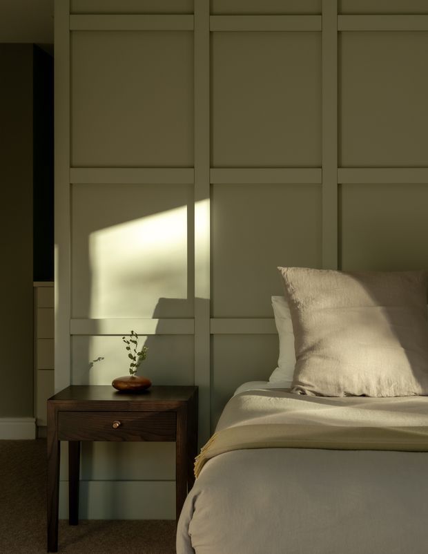 A warmly lit bedroom corner featuring a bed with cream bedding and textured pillows beside a dark wood bedside table. The wall behind the bed has a muted taupe-green painted grid of raised rectangular panel mouldings, while low late-afternoon sunlight creates a strong geometric highlight across the wall and bed. A small ceramic vessel with a sprig of greenery decorates the table, contributing to a calm, refined atmosphere.