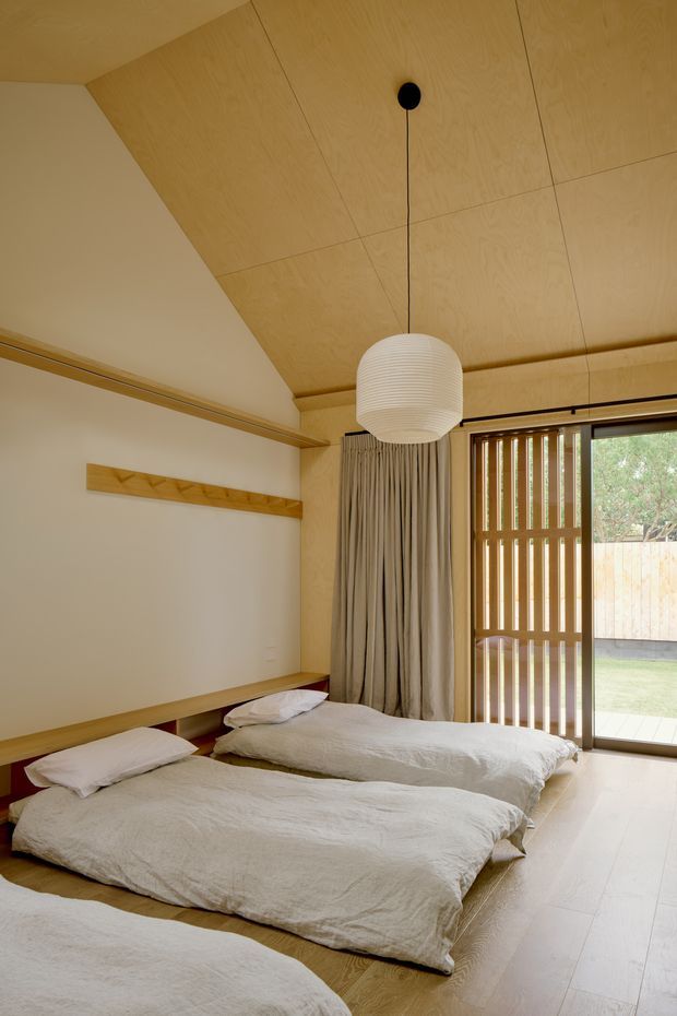 A warm, minimalist bedroom with multiple low-profile floor mattresses dressed in white linen. The room features extensive light timber ceiling panels and flooring, simple timber wall-mounted joinery, a sculptural white pendant light, full-height beige curtains, and timber vertical screening beside glazed sliding doors opening to a small outdoor area.