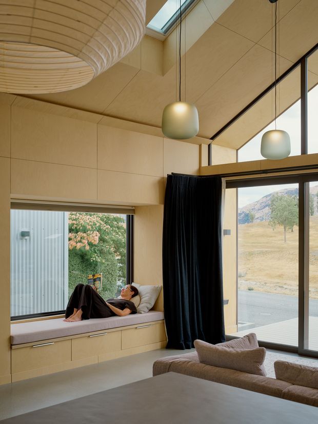 A warm, contemporary living room with extensive floor-to-ceiling glazing and a built-in window seat. Pale timber or plywood wall and ceiling panels create a continuous interior surface, complemented by dark curtains, sculptural pendant lights, and a large layered circular ceiling feature. A person relaxes on the upholstered window bench, while a sofa occupies the foreground. The room opens to a dry rural or mountainous landscape outside.