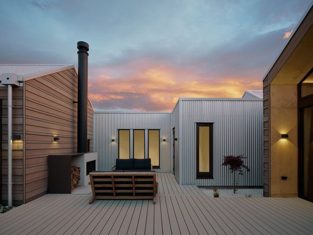 A contemporary courtyard-style outdoor living area enclosed by several modern structures. The space features a broad pale timber-look deck, horizontal timber cladding, corrugated metal wall and pitched roof forms, dark-framed windows and doors, built-in exterior lighting, a freestanding outdoor fireplace with chimney and stacked firewood, and a timber-framed lounge setting. The scene is photographed at dusk beneath a colourful sunset sky.