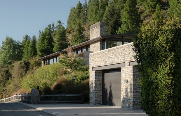 Exterior view of a contemporary hillside residence integrated into a densely wooded site. The building features pale stacked-stone masonry walls, dark metal-framed glazing, broad low-pitched roof planes, a prominent masonry chimney, and an integrated single-car garage. Landscaping and a driveway frame the foreground.