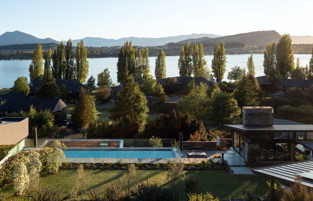 Wide elevated exterior view of a landscaped residential property beside a lake, with a long rectangular swimming pool in the foreground, a low modern pavilion with a flat roof at right, mature trees and gardens, and mountains across the water in the background.