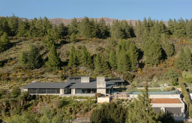 Wide exterior view of a low-profile contemporary house integrated into a steep, heavily planted hillside. The home has dark metal-clad pitched roof sections, extensive glazing, pale masonry or concrete chimney-like feature walls, terraces, and a visible swimming pool. Dense coniferous landscaping and rugged hills form the backdrop.