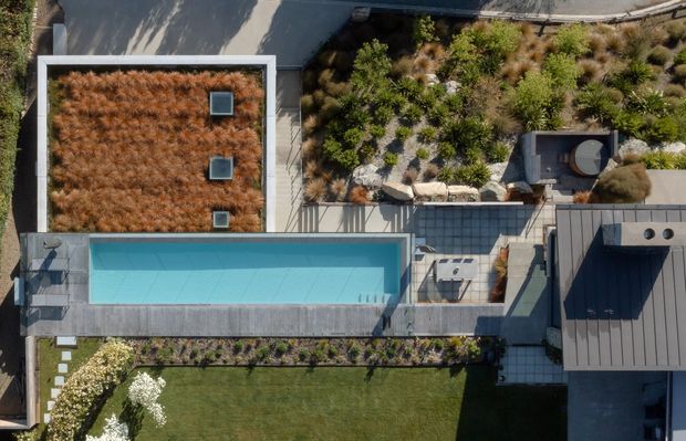 Aerial view of a contemporary residential property showing a long rectangular swimming pool, extensive landscaped roof or courtyard gardens with drought-tolerant planting, paved terraces, dark metal roofing, and a neatly maintained lawn. The composition emphasizes the site's integrated architecture, outdoor spaces, and planted surfaces.