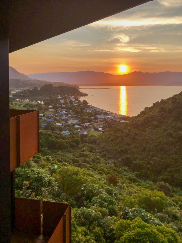 A dramatic sunset view from a hillside architectural lookout or balcony, overlooking a densely vegetated coastal or lakeside settlement, calm water, distant mountain ranges, and the sun reflected across the water. Rust-coloured solid panels and a broad overhead roof or canopy frame the view, suggesting an outdoor terrace or elevated dwelling integrated into the landscape.