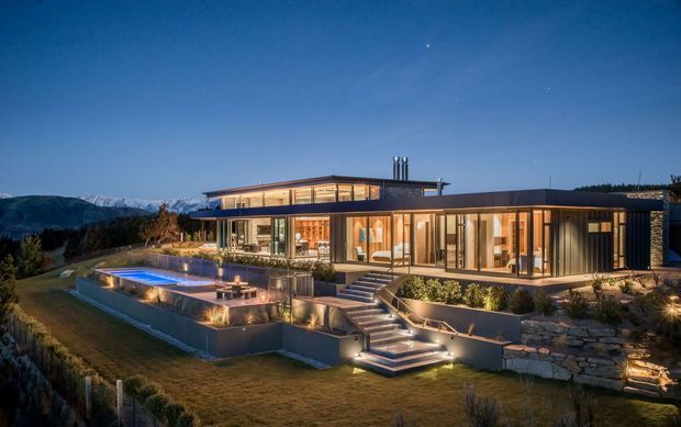 A dusk exterior view of a large contemporary single-storey residence on a landscaped, sloping site. The home features extensive floor-to-ceiling glazing, warm illuminated interiors, dark horizontal cladding, broad low-pitched roof forms, multiple terraces, and a raised swimming pool with illuminated steps and retaining walls. Snow-capped mountains are visible in the distant background.