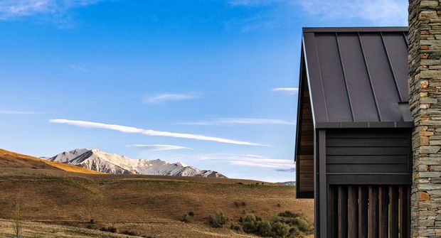 Exterior architectural detail of a contemporary mountain building, showing a dark standing-seam metal roof and cladding beside a rugged stacked-stone wall. The structure is set against an expansive alpine landscape with snow-dusted mountain peaks and open brown terrain.