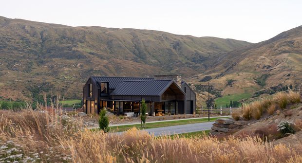 A contemporary dark-clad mountain house set within a landscaped rural site. The low, angular building has a prominent pitched metal roof, extensive glazed openings, timber-toned interiors visible through the windows, and a covered outdoor terrace. Dry ornamental grasses and stone landscaping frame the foreground, while steep rugged mountains dominate the background.