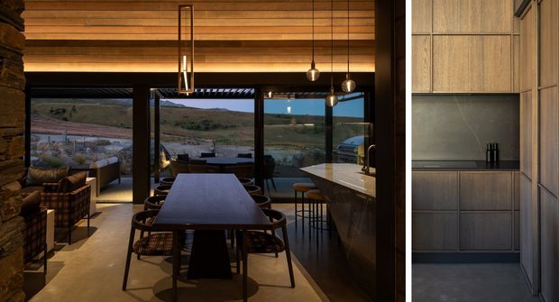 Warm, sophisticated open-plan dining and kitchen interior with extensive glazing overlooking a rugged landscape. The dining area features a long dark table with woven chairs, while the kitchen has dark wood cabinetry, a pale stone island, and a dark splashback. Timber ceiling beams, pendant lights, and a stone feature wall add texture and warmth. A narrow view at right shows additional built-in cabinetry and a tall integrated appliance or storage zone.