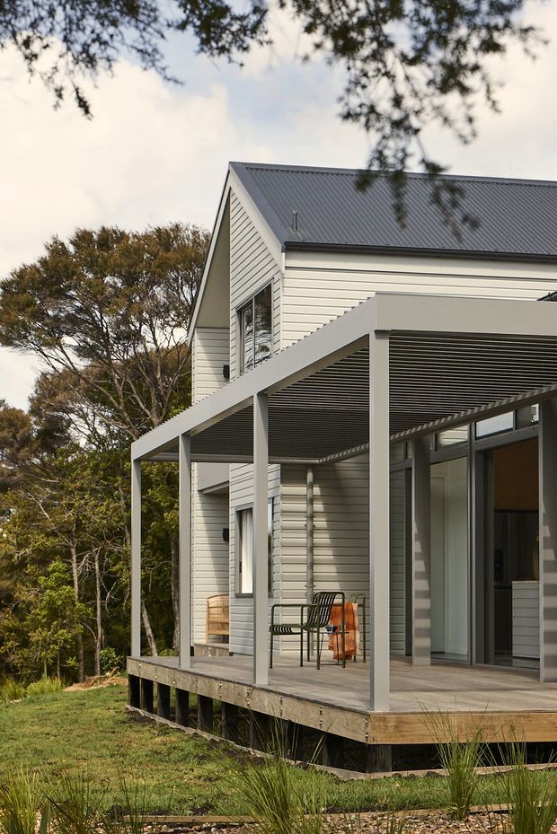 Exterior view of a contemporary timber-clad house set among established trees, featuring a pitched metal roof and a raised timber deck. A covered outdoor terrace runs along the facade, supported by simple square posts and fitted with horizontal louvres overhead. Large glazed doors open from the deck into the interior, with outdoor seating visible.