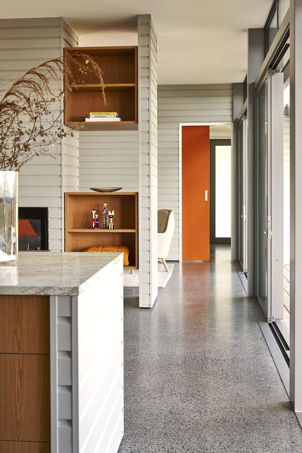 Standing beside the kitchen island, the view extends through the dining and living spaces towards the front entrance. An orange-painted cavity slider provides a playful burst of colour within the otherwise calm material palette.
