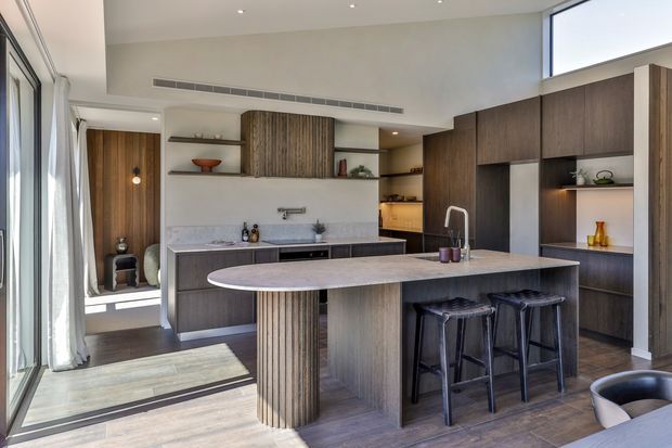 Contemporary open-plan kitchen with dark timber cabinetry, a large stone island with rounded ends, integrated shelving, a pale stone backsplash, and three bar stools. Full-height glazing and a glazed entry door bring in abundant natural light, while recessed ceiling lights and warm timber finishes create a refined, modern interior.