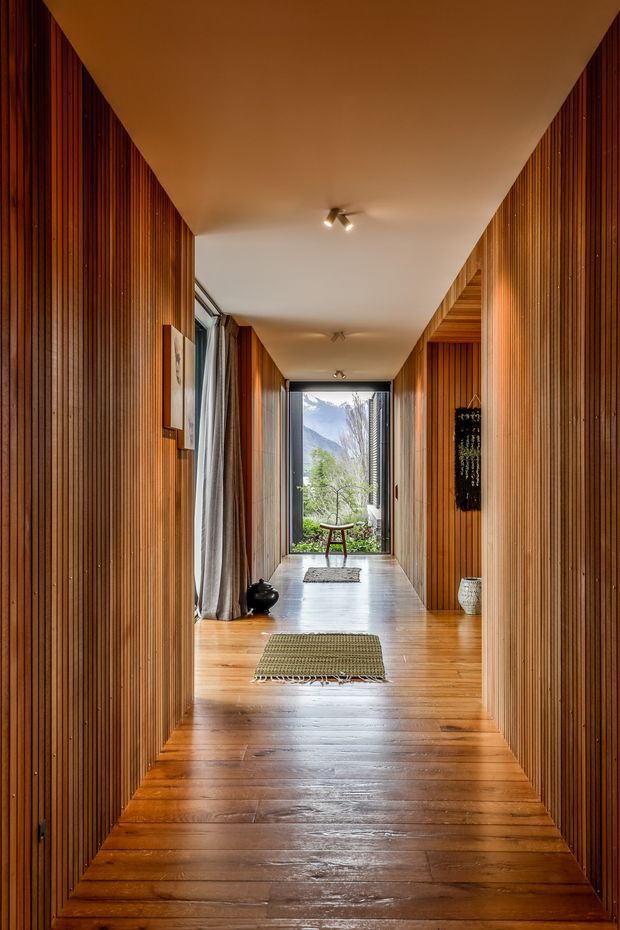 A long, narrow interior hallway lined with rich vertical timber slat wall panelling and matching timber flooring. The white ceiling has small recessed or surface-mounted spotlights, while full-height glazing at the far end frames a leafy mountain view. Curtains, small rugs, a stool, pottery, artwork and decorative wall pieces add warmth and texture. The space has a refined, warm timber-lined character with strong axial perspective.
