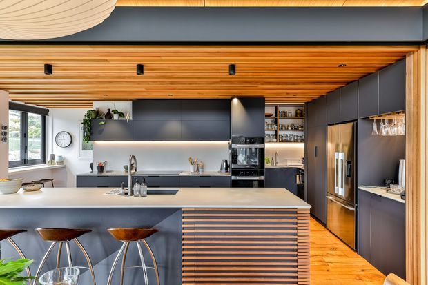 A sophisticated contemporary kitchen with a large pale stone island and integrated sink, dark charcoal cabinetry, built-in ovens, and a concealed refrigerator. Warm timber slats cover the ceiling and continue across the island front, complemented by timber flooring. Bar stools line the island, while open shelving with glassware and subtle under-cabinet lighting add detail. The space is brightened by large windows and indoor greenery, with a sculptural ceiling light partially visible overhead.