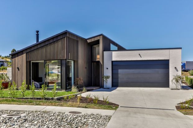 Contemporary single-storey house exterior with a dark vertically ribbed timber-clad living wing, recessed entry, extensive glazing, and a contrasting light rendered garage volume with a wide dark sectional garage door. The home sits on a landscaped suburban lot with minimalist planting and a concrete driveway.