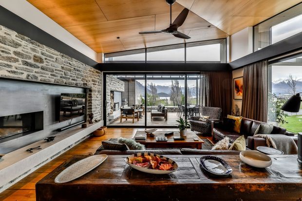 A spacious, contemporary living room with a dramatic floor-to-ceiling stone fireplace wall, warm timber-lined ceiling, dark-framed glazing, and an open connection to a dining area. The room contains layered sofas and armchairs, a substantial timber coffee table, and a large timber dining or console surface in the foreground. Expansive windows overlook a green landscape and mountains, while a ceiling fan and recessed lighting add practical features. The interior combines natural stone, timber, dark metal framing, and neutral furnishings in a refined lodge-like setting.