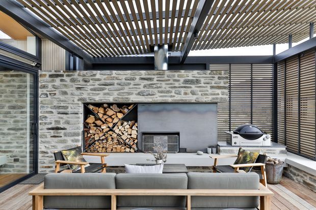 A refined outdoor living area with a covered timber pergola, exposed grey stone feature wall, built-in fireplace and firewood storage, barbecue, and a large contemporary outdoor lounge setting. Timber screening and glazed openings create a sheltered courtyard-like entertaining space.