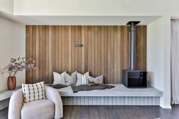 A refined living room featuring a built-in fireplace set into a vertical timber-panelled feature wall. A broad pale stone or concrete hearth bench runs beneath the fireplace and doubles as seating, layered with cushions and a throw. A curved upholstered armchair, ceramic vase with dried branches, dark timber flooring, and sheer curtains complete the calm, warm interior.
