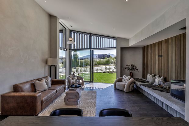Refined contemporary living room with a high ceiling, floor-to-ceiling glazing and vertical screening opening to a landscaped garden. The room features a large built-in timber feature wall and bench, a linear fireplace, brown leather seating, sculptural lounge chairs, a dark timber floor, and a dining or kitchen surface in the foreground. The neutral, earthy palette creates a warm, sophisticated atmosphere.