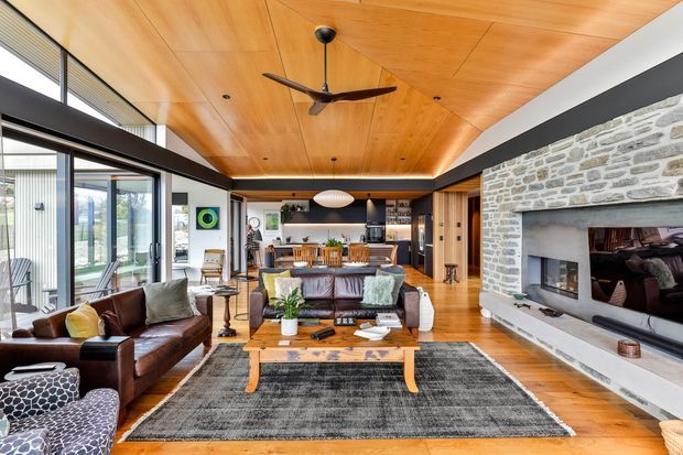 Open-plan living room with a warm timber-lined ceiling, exposed stone fireplace wall, polished timber flooring, large glazed sliding doors, leather seating, and a connected dining and kitchen area. A ceiling fan and contemporary furnishings create a relaxed, refined family living space.