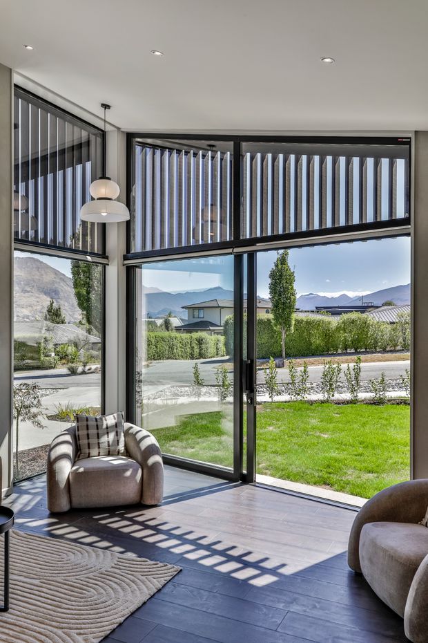 A bright, contemporary living room corner with expansive floor-to-ceiling black-framed glazing overlooking a landscaped suburban garden and mountain scenery. Vertical external sun-control fins span the upper windows, casting strong geometric shadows across the dark timber-look floor. The room includes rounded upholstered armchairs, a sculptural pendant light, and a textured neutral rug.