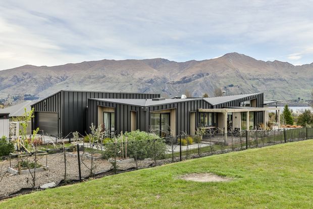 A contemporary single-storey residential building with a strongly articulated low-pitched roofline and dark vertical metal cladding. Large glazed openings and a partially covered outdoor terrace face a landscaped garden, with rugged mountains visible in the background. The property includes fenced planting beds, lawn, and a water body or valley landscape beyond.