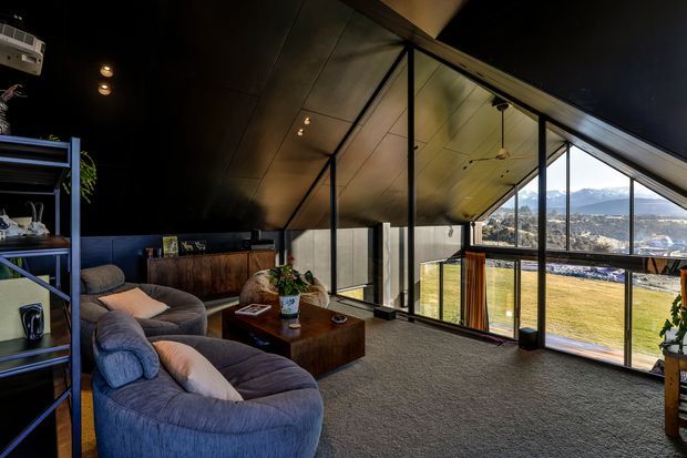 A moody, loft-like living room with a large curved sectional sofa, dark coffee table, open shelving, and a dramatic sloping ceiling. Floor-to-ceiling glazing frames views of a grassy site and distant mountains, while black structural framing and warm timber ceiling panels create a contemporary retreat atmosphere.