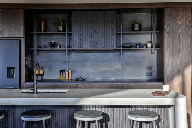 A dark, sophisticated contemporary kitchen with a long stone island and three bar stools in the foreground. The kitchen features dark vertically grained timber cabinetry, open shelving with black metal framing, a large integrated refrigerator, a grey tiled or slab backsplash, stainless steel sink and tapware, and understated decorative objects. The composition emphasizes the island, cabinetry, and bar seating, with a moody, refined material palette.