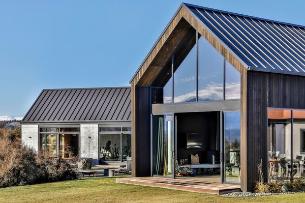 Exterior architectural photograph of a contemporary two-part residence with steep pitched gabled roofs, dark standing-seam metal roofing, vertical dark timber cladding, exposed timber soffits, and expansive floor-to-ceiling glazing. A glazed living area and outdoor landscaping are visible in the foreground, with a lower wing extending behind.