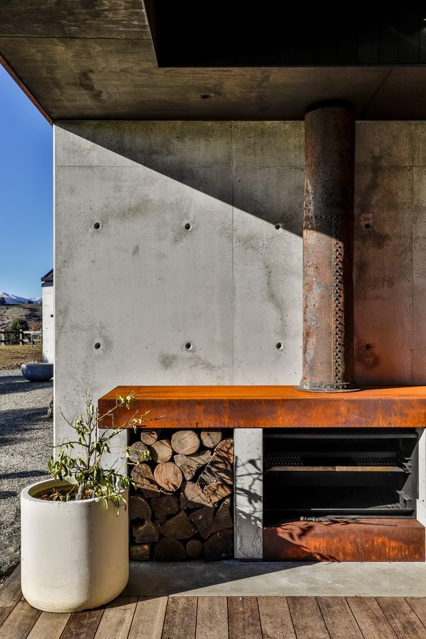 Close-up exterior view of a built-in outdoor barbecue and fireplace installation beneath a deep concrete overhang. The composition features a raw concrete wall with exposed fixing points, a weathered rusted steel chimney and counter, integrated firewood storage, and a dark built-in grill. Timber decking, a large planter, and a distant rugged landscape are visible around the structure. The design has a rugged, industrial character with strong sunlight and material contrast.