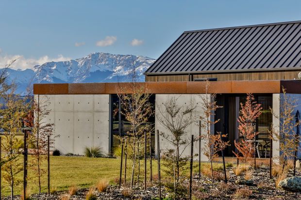Contemporary single-storey home set against a dramatic snow-capped mountain backdrop. The building combines a steep dark standing-seam metal gable roof with smooth concrete wall panels, extensive dark-framed glazing, and a rust-toned horizontal metal band. A landscaped garden with young trees and grasses surrounds the residence.