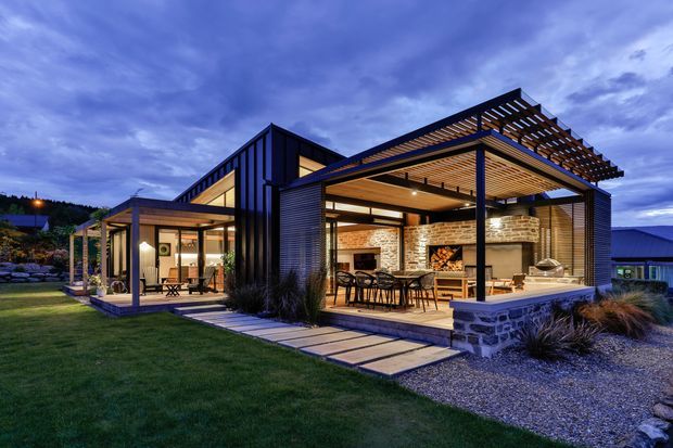 Contemporary single-storey home photographed at dusk, featuring a dark vertical-clad central volume, extensive glazing, and a large covered outdoor entertaining pavilion. The pavilion has a timber-lined ceiling, black steel framing, exposed brick feature walls, an outdoor fireplace, dining and lounge areas, and stone-clad base walls. Paved stepping stones connect the covered terrace to the landscaped lawn and gravel surrounds.