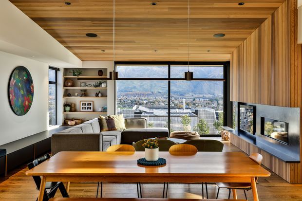 A warm, contemporary open-plan living and dining area with expansive mountain and city views through floor-to-ceiling glazing. The room features a large timber dining table, upholstered dining chairs, a sectional sofa, built-in shelving, a wall-mounted fireplace set into vertical timber panelling, and a timber-lined ceiling with recessed lighting.