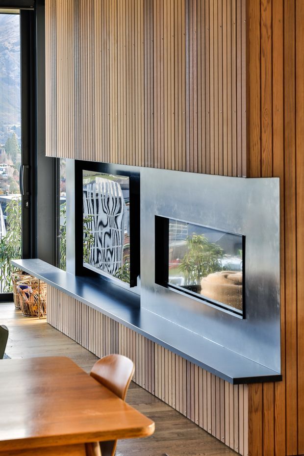 Interior view of a contemporary dining area featuring a full-height vertical timber slat wall and built-in dark metal display/fireplace-style surround with inset reflective openings. A timber dining table and chair sit in the foreground, while large windows provide views to the surrounding landscape. The space has warm wood tones balanced by charcoal and metallic grey surfaces.