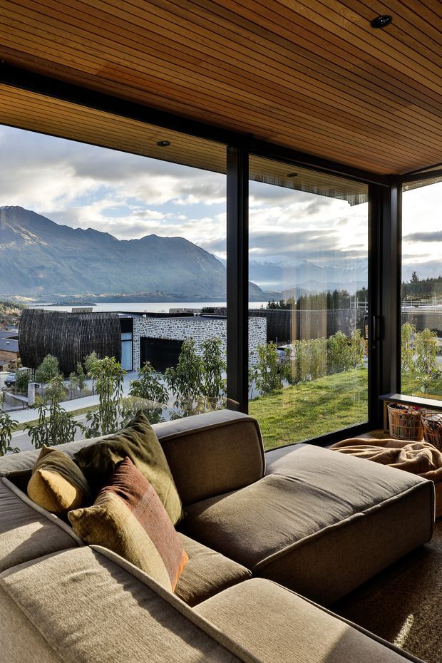 A warmly lit living room with a large taupe sectional sofa positioned beside floor-to-ceiling windows. The room features a striking slatted timber ceiling, dark-framed glazing, and expansive views of a lake and mountainous landscape. Soft cushions and throws add a relaxed, inviting feel, while the exterior garden and neighbouring structures are visible beyond the glass.