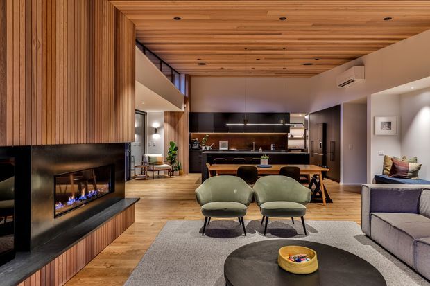 A warm, open-plan contemporary living area with a timber-lined ceiling and feature wall, a built-in linear fireplace, lounge seating, occasional chairs, and a kitchen with island bench in the background. The space uses timber flooring, dark joinery, soft neutral furnishings, and recessed lighting.
