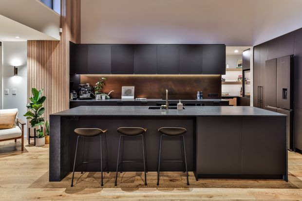 A contemporary open-plan kitchen viewed straight on, dominated by a large dark island with three bar stools. The kitchen features matte dark cabinetry, a warm timber-toned splashback, integrated appliances, a stainless or dark metal sink mixer, and light timber flooring. Vertical timber wall panelling and glimpses of adjacent living and circulation areas are visible at the sides.