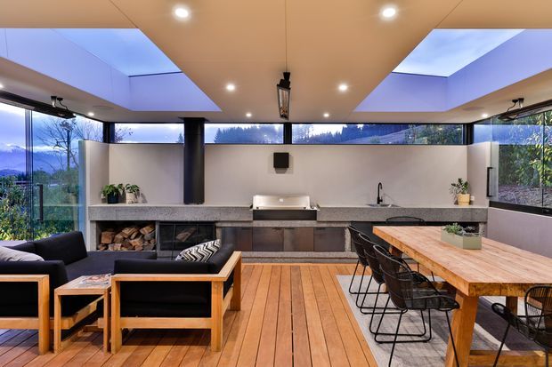 A contemporary open-plan indoor-outdoor entertaining space with a kitchen, dining area, and lounge. The room features extensive corner glazing, a timber-lined ceiling with recessed lighting, warm timber flooring, dark-framed windows, a long stone kitchen bench with integrated cabinetry, a wood-burning fireplace niche, and a large timber dining table with black wire-frame chairs. Mountainous landscape is visible through the glazing.