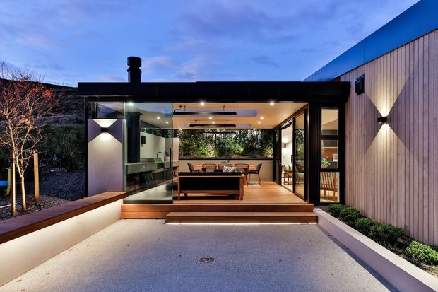 A contemporary single-storey home viewed from an illuminated paved courtyard at dusk. The building combines a dark, flat-roofed glazed pavilion with a timber-clad side wall, broad sliding glass openings, exterior steps, integrated wall lighting, and a sheltered outdoor seating area. A living room is visible through the glazing, with a landscaped courtyard and built-in bench framing the approach.