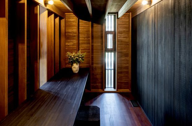 A dark, narrow timber-lined interior with a long built-in bench or console along the left wall, vertical wood-panelled walls, a slim clerestory-style window, and exposed structural ceiling beams. Warm recessed and pendant-like lighting creates a dramatic, intimate atmosphere. The space appears to be a hallway or entry passage, with a small potted plant placed on the bench.