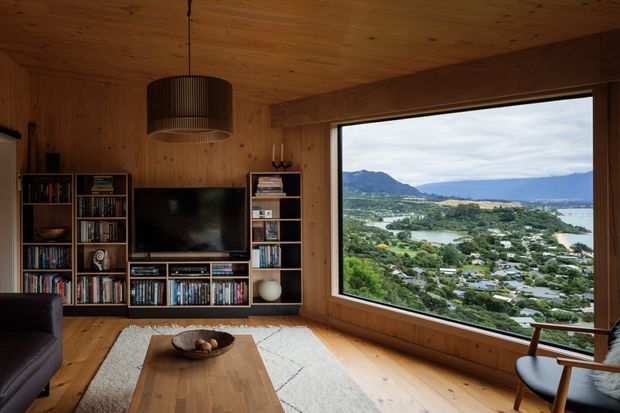 A warm, wood-lined living room with a built-in media and bookcase wall, television, pendant light, timber coffee table, rug, and lounge seating. A large picture window provides an expansive view across a green residential landscape, water, and mountains. The room has a cosy, natural, cabin-like atmosphere with strong timber detailing.