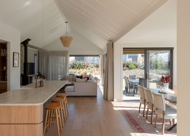 Bright open-plan interior combining a kitchen, dining area and living room beneath a high pitched, white-lined ceiling. The kitchen features a large light stone island with timber cabinetry and timber stools. A dining table sits beside expansive glazed doors opening to an outdoor patio, while the living area contains a pale sectional sofa and a dark fireplace or media unit. Warm timber flooring, neutral furnishings, sheer curtains and a woven pendant create a relaxed, refined atmosphere.