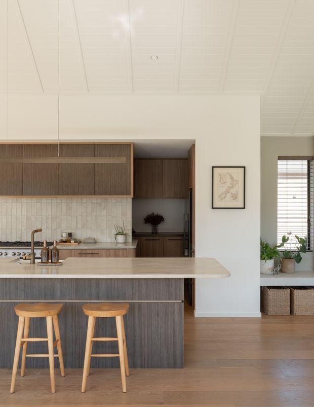 Bright, refined kitchen interior with a large island and two timber stools. The kitchen features dark timber-grain cabinetry, a pale stone benchtop, light rectangular tiled splashback, integrated pantry joinery, and warm timber flooring. A white vertically panelled ceiling, framed artwork, indoor plants, woven baskets, and a window with timber blinds complete the calm, natural scheme.