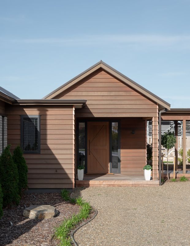 Front exterior view of a single-storey house with warm horizontal timber weatherboard cladding, a prominent gabled roof, covered entry porch, timber front door with sidelights, and simple landscaped gravel approach.