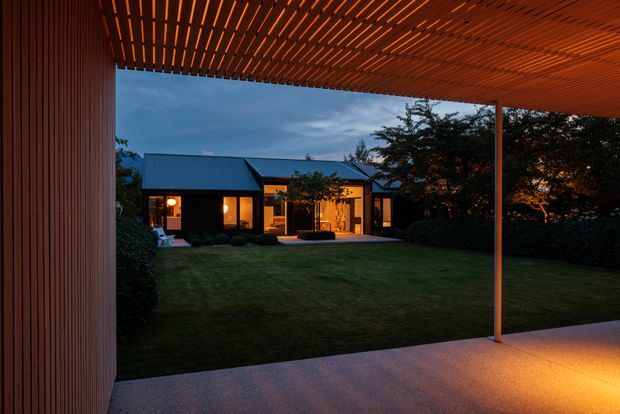 Dusk view from a covered outdoor terrace or patio toward a single-storey contemporary home. The foreground features a warm timber slatted pergola ceiling, slender support post, pale concrete floor, and a broad manicured lawn leading to the dark-clad house. Warm interior and exterior lighting glows through the windows, creating a sheltered and inviting connection between the terrace and garden.