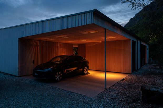 A dramatic dusk exterior of a contemporary single-storey garage and carport structure. The building has a simple gabled roof form, dark blue-grey corrugated metal cladding, and a deep covered parking bay supported by a slender post. Warm concealed lighting illuminates the timber-lined ceiling and vertically ribbed timber wall surfaces around a parked car. The structure sits against a dark, steeply wooded hillside on a gravel forecourt.