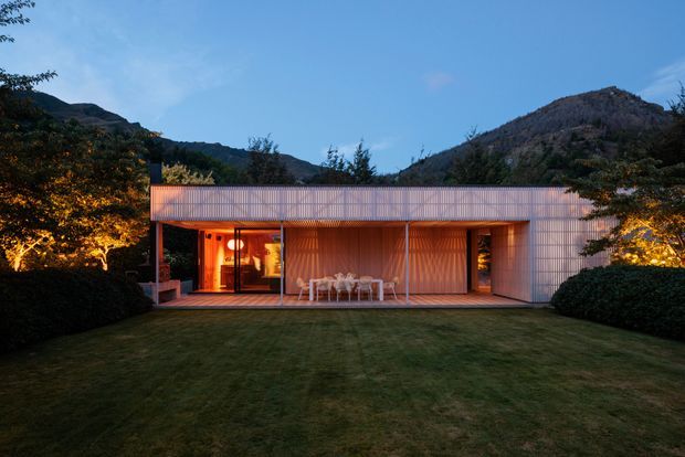 A low, minimalist pavilion-like house set against wooded mountain slopes at dusk. The building has a long horizontal form with a flat roof, a translucent or finely screened pale facade, extensive open glazing, and a covered outdoor terrace furnished with a dining table and chairs. Warm interior lighting contrasts with the cool blue evening sky and dark landscaped lawn.